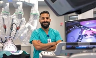 Surgeon in teal scrubs standing with arms crossed in operating room with robotic surgical equipment and monitor
